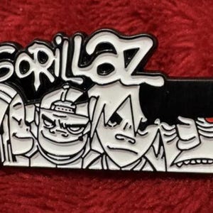 May include: A black and white enamel pin featuring the word "Gorillaz" in a stylized font. The pin showcases the band's cartoon characters in a rectangular design. The pin is set against a red background.