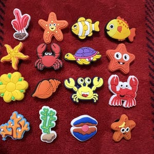 May include: A collection of colorful, cartoon-style sea creature charms, including starfish, crabs, fish, and coral. The charms are in various colors like orange, yellow, red, and blue, and are arranged on a red textured surface.
