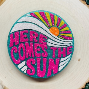 May include: A teal blue round wooden sign with a pink glitter design that says "Here Comes The Sun" with a yellow and orange glitter sun rising over a white glitter wave.
