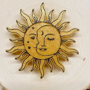 May include: A golden sun and moon design with black outlines. The sun has radiating rays and a smiling face, while the moon has a crescent and a closed-eye face. The celestial design is set against a light wood background.