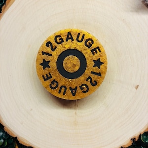May include: A gold-colored, circular object with the text "12 GAUGE" in black. The object has a black center circle and two black star shapes. The item is set on a light brown, wooden surface.