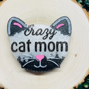 May include: A cat-shaped decoration with the words "crazy cat mom" in black script. The cat's face is gray and white with pink accents on the ears and nose. The decoration is set on a wooden slice.
