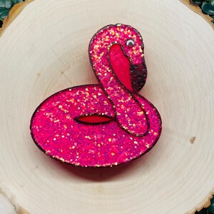 May include: A pink glitter flamingo pool float with a white eye. The float is on a wooden surface.