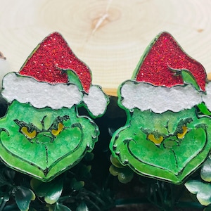May include: Two green Grinch-themed Christmas ornaments. Each ornament features the Grinch's face with a red glitter Santa hat trimmed with white faux fur. The ornaments are set against a backdrop of greenery and a wooden slice.