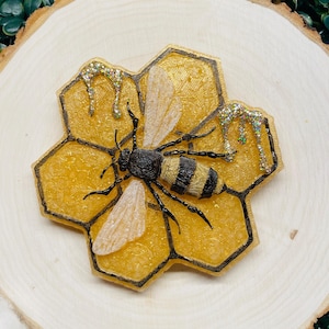 May include: A resin bee with black and yellow stripes is perched on a yellow honeycomb with black outlines. The honeycomb has gold glitter dripping from the top.