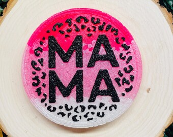 May include: A round coaster with the word "MAMA" in black glitter letters. The coaster has a pink and white ombre design with black leopard print accents. The coaster is displayed on a wooden slice.