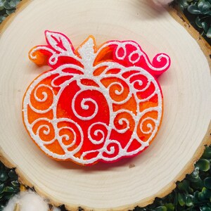May include: A decorative pumpkin in shades of orange and red, with white swirling accents. The pumpkin is placed on a wooden slice, with greenery and cotton accents in the background. A festive fall decoration.