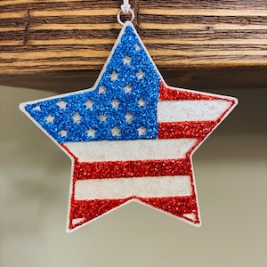May include: A star-shaped ornament with a glittery American flag design. The blue section features white stars, while the stripes alternate between red and white glitter. It hangs from a white string.