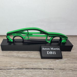 May include: A green Aston Martin DB11 car outline on a black base. The car's design is layered, creating a 3D effect. A black plaque with "Aston Martin DB11" is in front of the car. The background is a light colour.