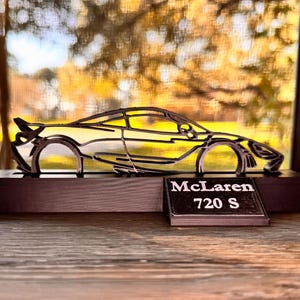 May include: A black metal silhouette of a McLaren 720S car on a black wooden base. The car is displayed against a blurred background of trees and foliage.