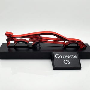Könnte beinhalten: Rote Corvette C8 Autoskulptur. Das Auto ist in einem stilisierten Open-Frame-Design dargestellt und auf einem schwarzen rechteckigen Sockel montiert. Ein kleines schwarzes Schild zeigt den Text "Corvette C8".