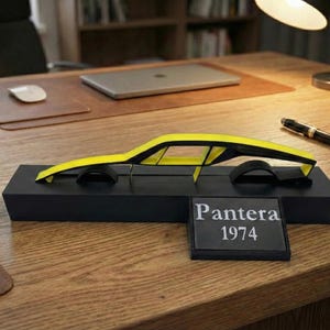 May include: A yellow and black model car with the text "Pantera 1974" on a black base. The car is a sleek, modern design, and sits on a wooden desk.