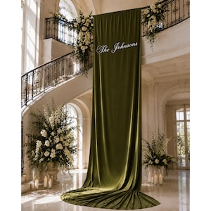 May include: A long, floor-length olive green velvet fabric backdrop featuring the white cursive text The Johnsons centered near the top. The fabric hangs vertically from a high point, creating soft folds that pool onto the floor.