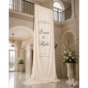 May include: A long, cream-colored fabric banner hanging vertically, featuring black printed text that reads: Welcome to the wedding of Evan & Rylie, La Savoie, February 1, 2026. The banner is draped from an upper balcony down to the floor.