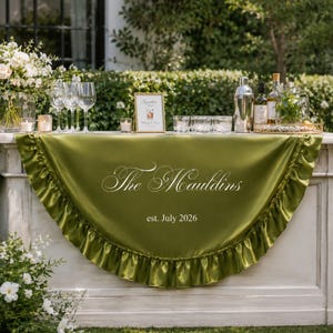 May include: A semi-circular olive green satin table runner with a ruffled edge, featuring the white embroidered text "The Mauldins" and "est. July 2026" centered on the fabric, draped over the front of a white rectangular bar counter.
