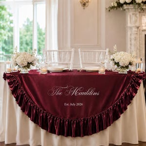 May include: A deep burgundy satin table skirt featuring a ruffled edge and white embroidered text that reads The Mauldins and Est. July 2026. The fabric is draped over a white tablecloth, creating a formal display for a two-person table setting.