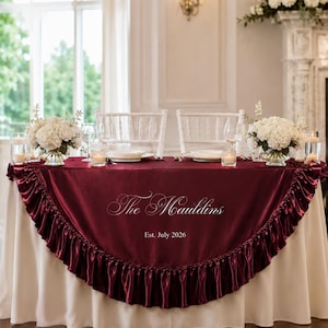 May include: A burgundy satin table skirt with a ruffled edge and a white base tablecloth. The fabric features the white embroidered text "The Mauldins" and "Est. July 2026" centred on the front. Two white chairs and floral centrepieces are visible.
