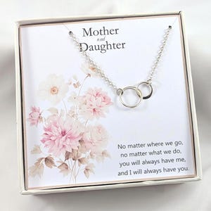 May include: A silver necklace with two interlocking circles, presented in a gift box. The box features the words "Mother and Daughter" and a floral design with pink and white flowers. The text on the box reads, "No matter where we go..."