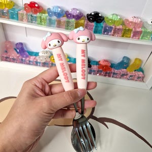 May include: A pink My Melody themed children's fork and spoon set with a pink handle and stainless steel utensils.