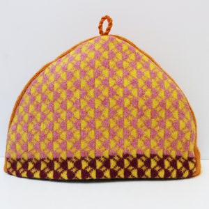 May include: A knitted tea cosy in a triangular shape. The cosy features a repeating pattern of yellow and pink squares, with a dark burgundy border at the bottom. It has an orange trim and a small loop at the top.