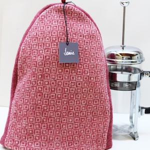 May include: A dome-shaped tea cozy featuring a repeating geometric square pattern in shades of red and light pink. A small dark gray square tag with the white text Janie is attached to the top loop. A glass and chrome French press sits beside it.