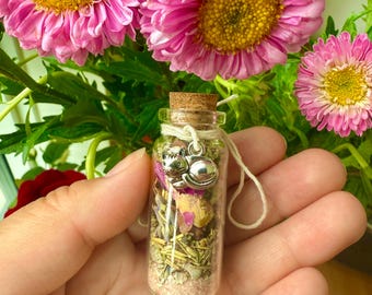 May include: A small glass bottle with a cork stopper, filled with dried herbs and pink sand. A silver cat charm hangs from the bottle. The bottle is surrounded by pink flowers with yellow centres.