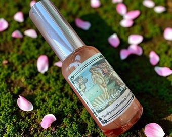 May include: A brown glass spray bottle with a silver cap, labeled "Aphrodite Touch Mist." The label features a Renaissance painting and text. The bottle rests on green moss, surrounded by pink flower petals.