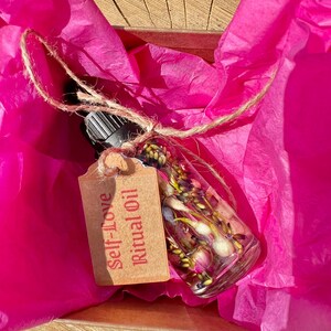 May include: A small glass bottle filled with dried flowers and a brown paper tag that reads "Self-Love Ritual Oil" is nestled in pink tissue paper.