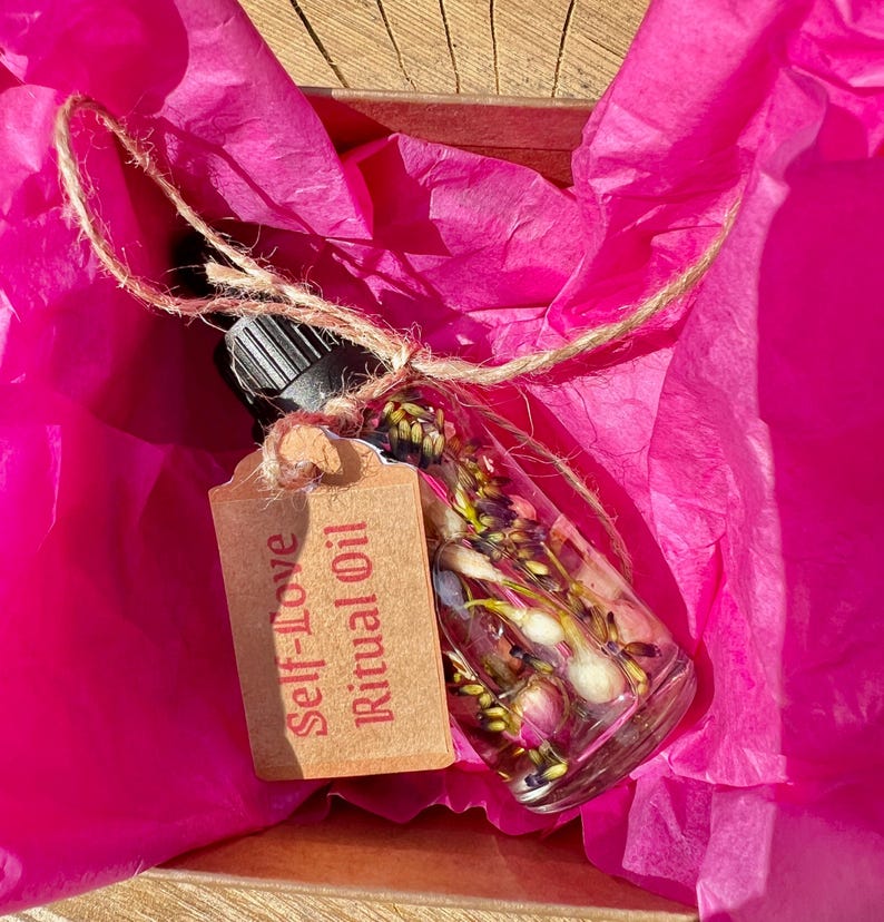 May include: A small glass bottle filled with dried flowers and a brown paper tag that reads "Self-Love Ritual Oil" is nestled in pink tissue paper.