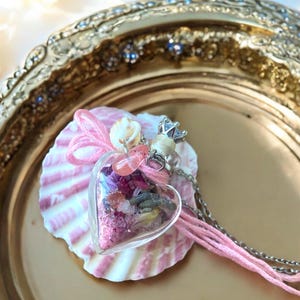 May include: A heart-shaped glass pendant necklace filled with dried flowers and crystals. The pendant hangs from a silver chain and rests on a pink and white seashell. The necklace is decorated with pink string and small seashells.