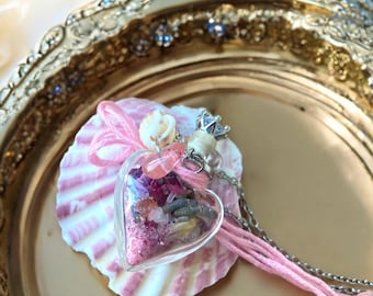 May include: A heart-shaped glass pendant necklace filled with dried flowers and crystals. The pendant is suspended from a silver chain and rests on a pink and white seashell. The necklace is adorned with pink string and small seashells.