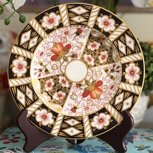 May include: Decorative plate with a floral design. The plate features a white center with a circular indentation, surrounded by sections of red, gold, and black patterns. The rim has alternating black and gold geometric shapes and floral motifs.