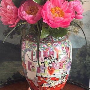 May include: A vibrant floral arrangement of pink peonies in a decorative, hand-painted porcelain vase. The vase features a detailed, colorful scene with figures and intricate patterns. The lid of the vase is visible in the foreground.