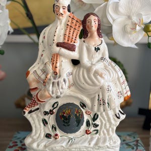 May include: A white ceramic figurine depicting a seated couple in traditional attire. The man holds a musical instrument and wears a hat. The woman is in a white dress. The base features a floral design and a central emblem. The figurine has a vintage aesthetic.