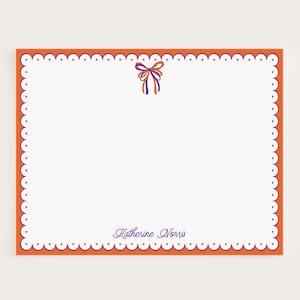 May include: A white stationery card with an orange border and a scalloped edge. A small bow in purple, orange, and blue is at the top. The name "Katherine Norris" is written in purple script at the bottom.