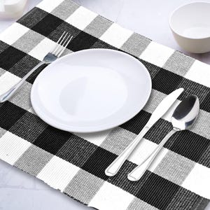 May include: A white plate, bowl, and silverware set on a black and white checkered placemat. The silverware includes a fork, knife, and spoon. The placemat has a classic gingham pattern.