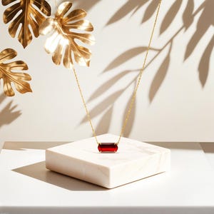 May include: A gold chain necklace with a red rectangular pendant on a white marble surface. The necklace is surrounded by gold leaf.