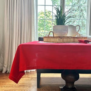 May include: A vibrant red tablecloth covers a dark wooden table. A woven tray holds a potted plant and a white pitcher. The scene is set in a well-lit room with a large window and white curtains, creating a warm and inviting atmosphere.