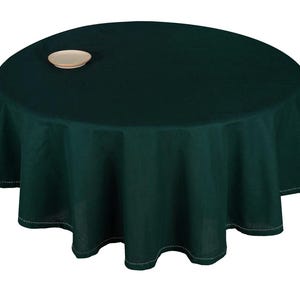 May include: A dark green round tablecloth with a scalloped edge and white stitching. A small, round, beige plate sits on the tablecloth. The tablecloth is made of a solid, opaque fabric.