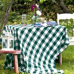 May include: A green and white checkered tablecloth is laid out on a wooden table in a garden setting. A white lantern, a glass jar, and a bouquet of flowers are on the table. A white chair is in the background.