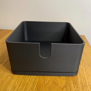 May include: A black square-shaped storage box with a slot in the front. The box is made of a smooth, matt material.