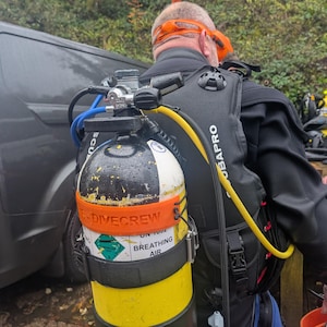 May include: A black scuba diving tank with a yellow and black band around it. The tank has a black and orange strap with the text "Dive Crew" on it. The tank is attached to a black scuba diving backpack with a yellow hose.