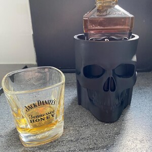 May include: A black skull-shaped holder containing a Jack Daniel's bottle, and a Jack Daniel's Tennessee Honey shot glass. The shot glass is filled with a golden liquid. The bottle is labelled "Old No. 7 Brand".