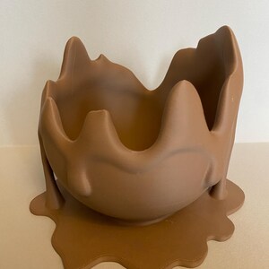 May include: A brown ceramic planter shaped like a melting chocolate egg. The planter has a wide opening at the top and a base that is shaped like a puddle of melted chocolate.