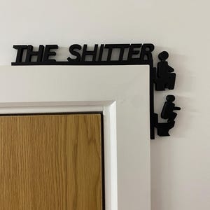 以下が含まれることがあります： 「THE SHITTER」と書かれた黒い木製の看板と、トイレの個室にいる2人の棒人間。