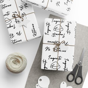 May include: White gift wrap with black text that says "Ingrate" and "No Returns". The text is written in a handwritten style. There is a small illustration of a Santa hat on the wrapping paper.