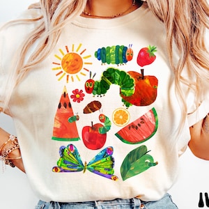 Puede incluir: Camiseta de color marfil con una colorida ilustraci&oacute;n de una oruga, un sol, frutas y una mariposa. El dise&ntilde;o incluye una oruga verde, una manzana roja y una rodaja de sand&iacute;a. La palabra "IVORY" est&aacute; impresa en negro en la parte inferior.