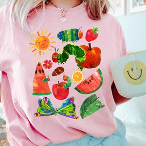 Puede incluir: Sudadera rosa claro con un dise&ntilde;o colorido de una oruga, frutas y una mariposa. El dise&ntilde;o incluye un sol, una manzana, una sand&iacute;a y una rodaja de naranja. La obra de arte est&aacute; inspirada en un libro infantil.