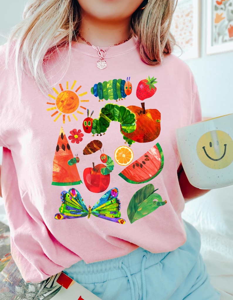 Puede incluir: Sudadera rosa claro con un dise&ntilde;o colorido de una oruga, frutas y una mariposa. El dise&ntilde;o incluye un sol, una manzana, una sand&iacute;a y una rodaja de naranja. La obra de arte est&aacute; inspirada en un libro infantil.