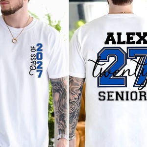 Może przedstawiać: Biały t-shirt z kr&oacute;tkim rękawem z niebieską grafiką pionową na piersi z napisem "CLASS OF 2027". Z tyłu znajduje się imię "ALEX" w kolorze czarnym, liczba "27" w kolorze niebieskim, słowo "twenty" pisane kursywą oraz "SENIOR" w kolorze czarnym.